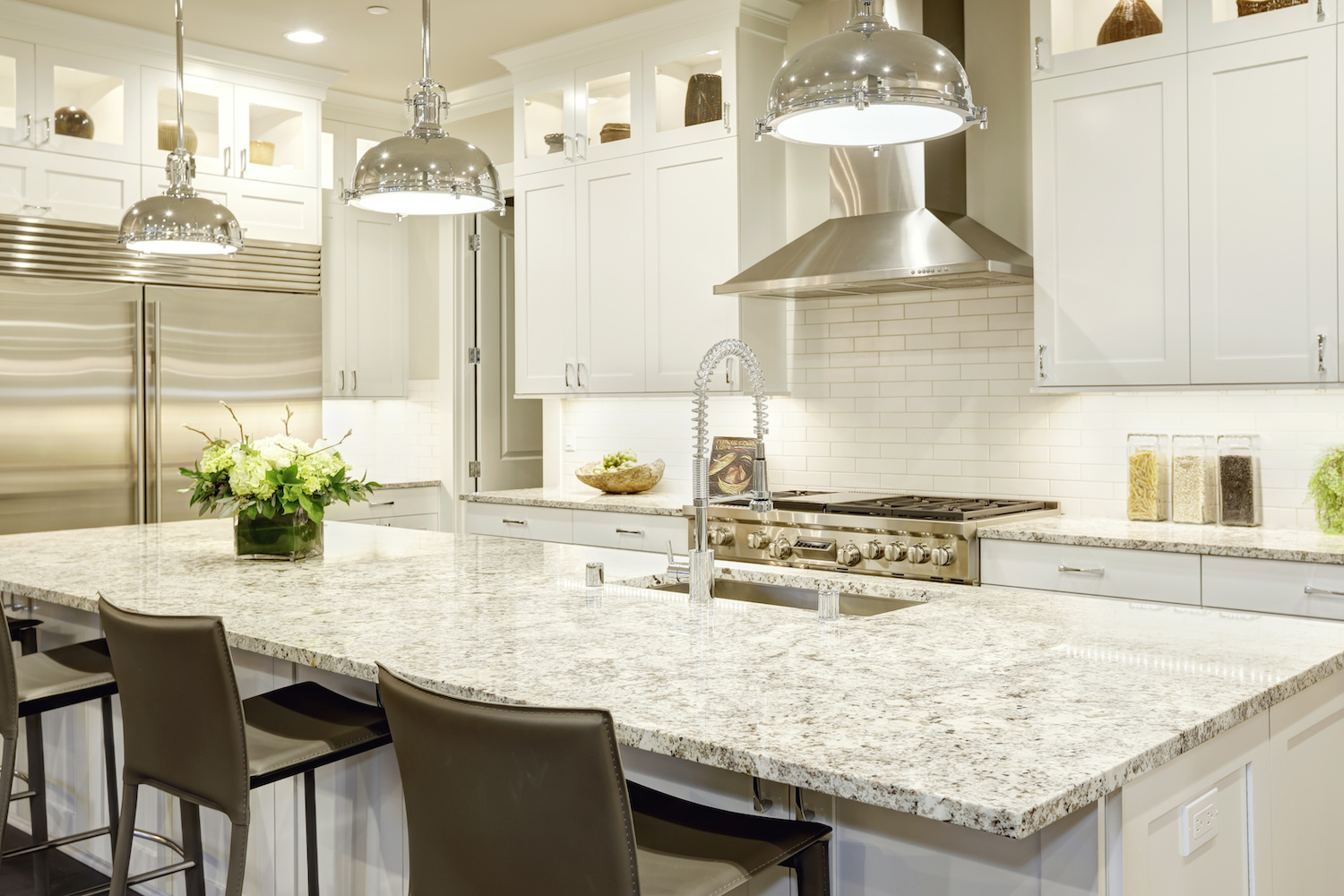 White kitchen design features large bar style kitchen island with granite countertop illuminated by modern pendant lights. Stainless steel appliances framed by white shaker cabinets . Northwest, USA